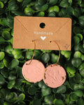 A pair of handmade round dangle earrings with gold-tone hoops and dusty pink circular charms featuring embossed floral designs and distressed gold accents, displayed on a brown kraft card over a green leaf background.