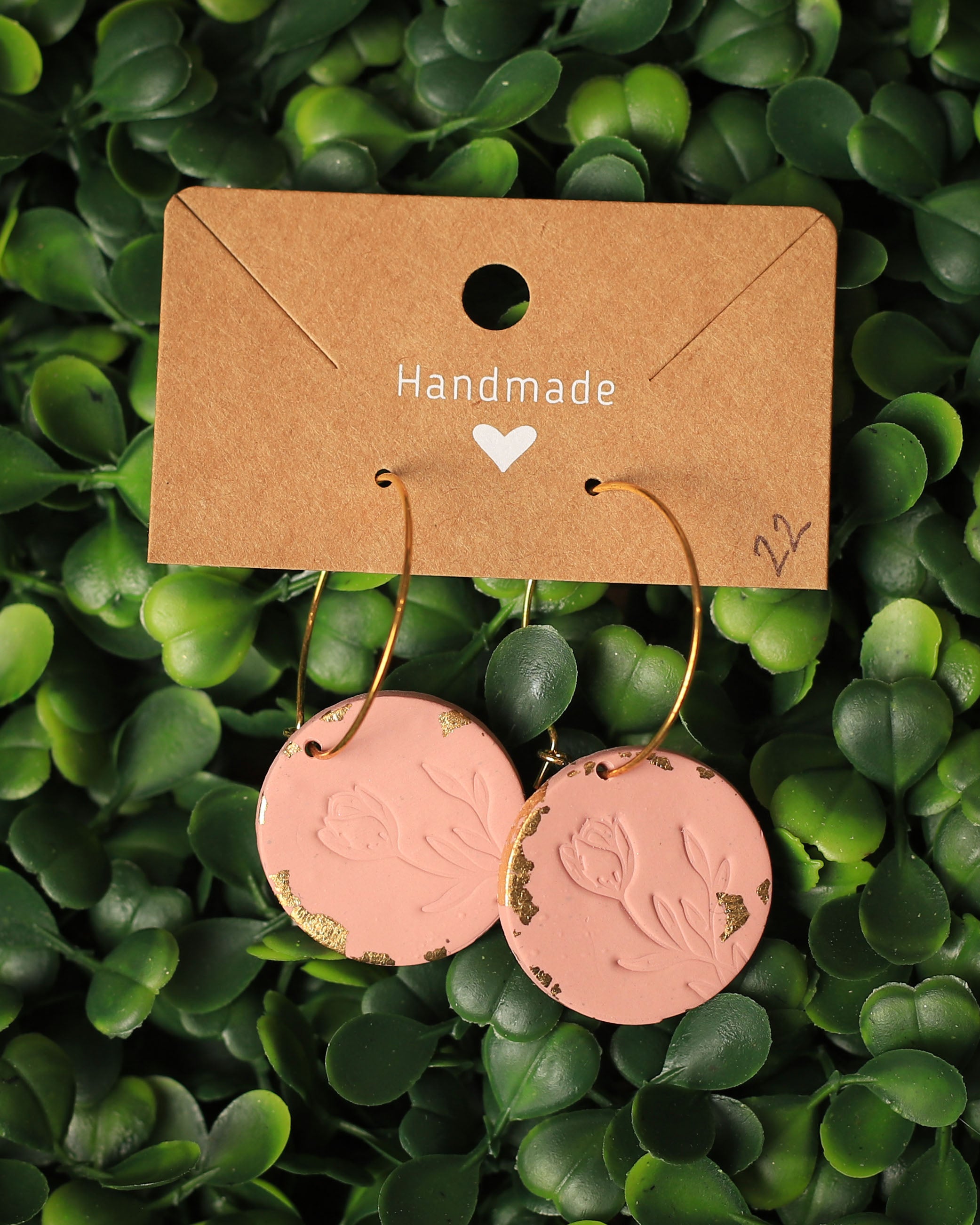 A pair of handmade round dangle earrings with gold-tone hoops and dusty pink circular charms featuring embossed floral designs and distressed gold accents, displayed on a brown kraft card over a green leaf background.